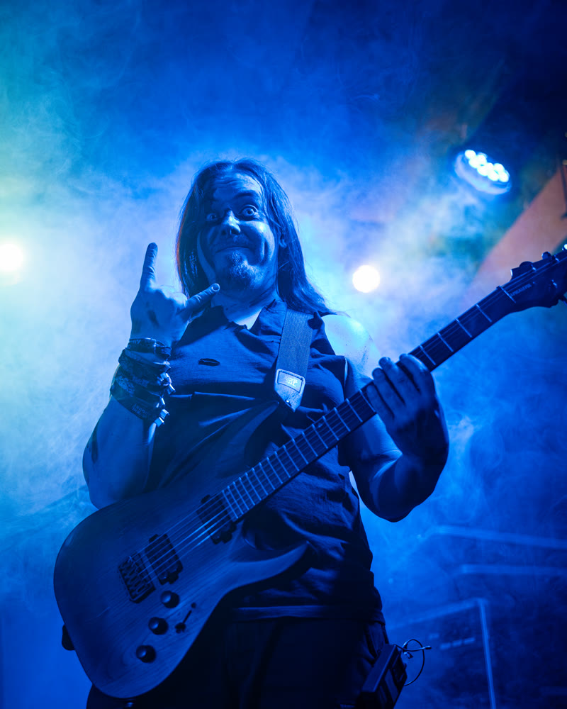 Riese on stage in blue light, holding his guitar, his free hand raised in the horns