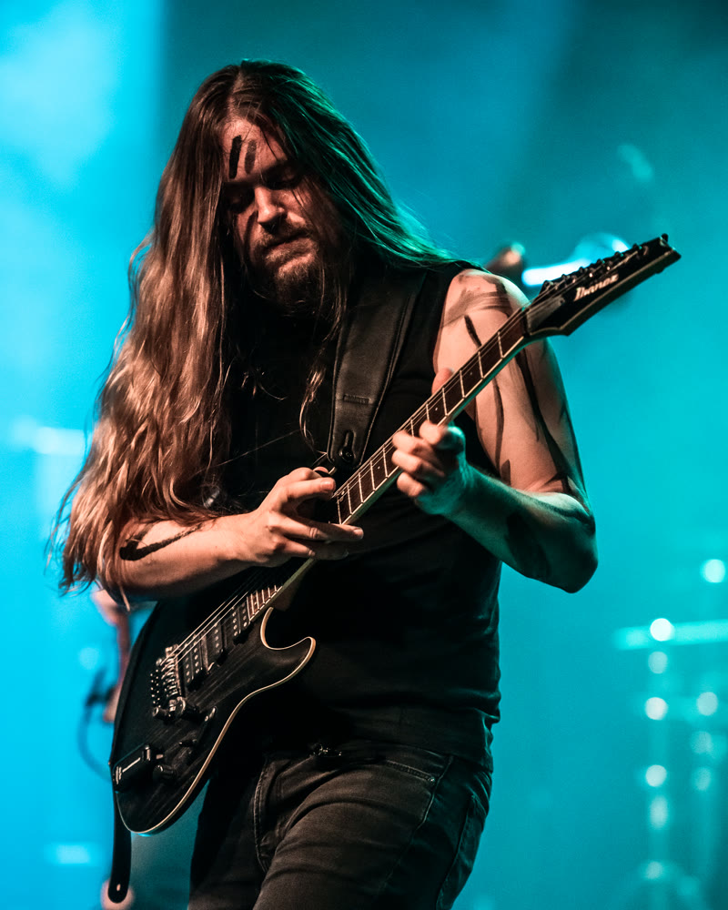 Thomas in turquoise stage light, bent over his guitar, long hair across his face